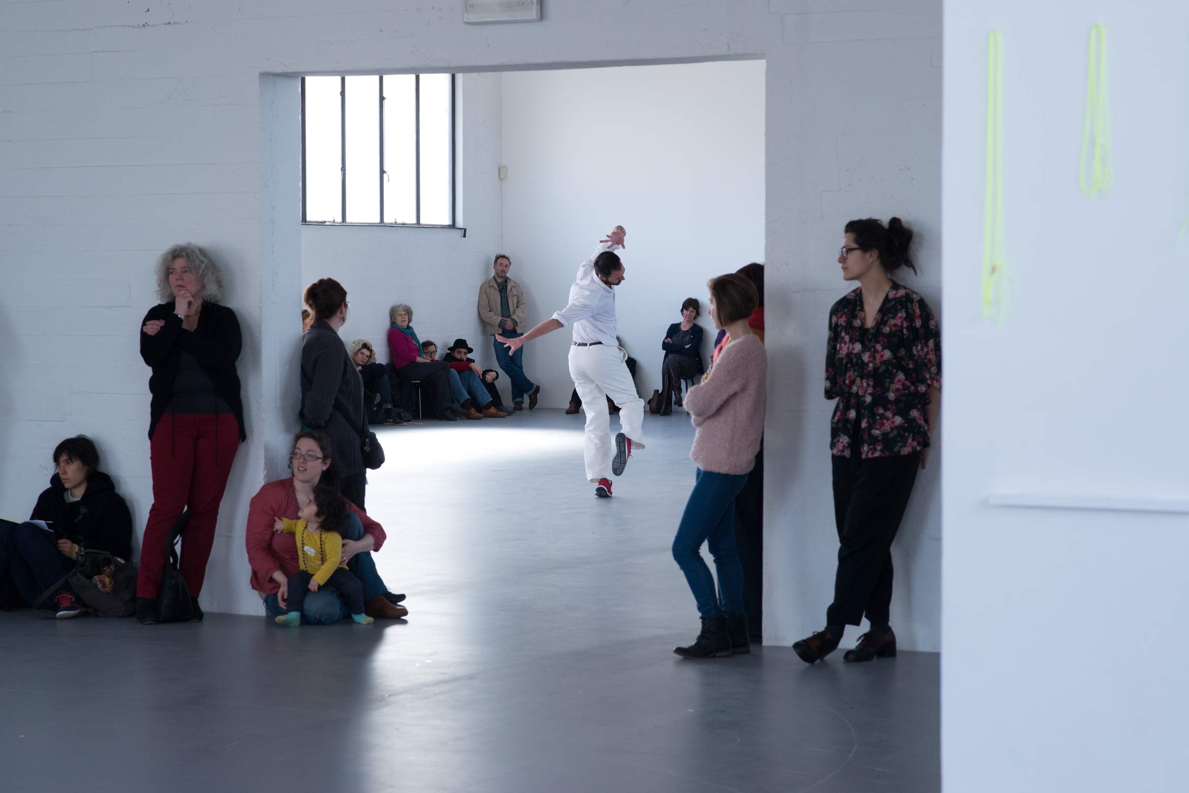 A dancer in a twisted posture is framed by an opening between two museum spaces, with visitors gathered around in both rooms observing.