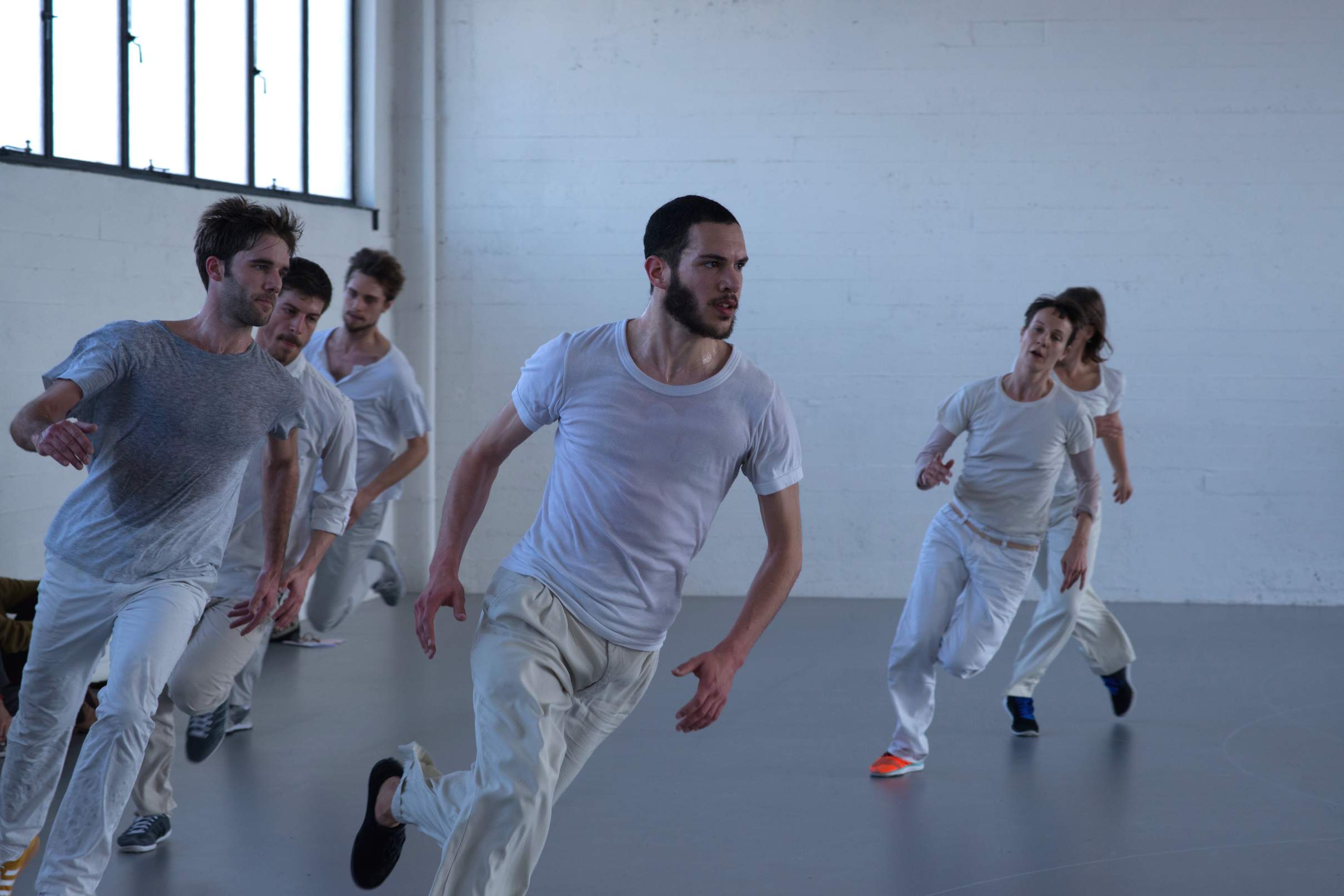 Six dancers run along a curved path in a white museum space, moving with high energy.