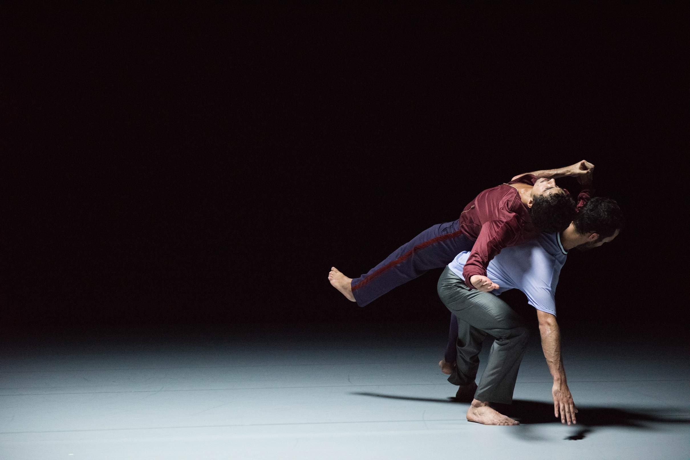 A dancer supports another in a careful balance, backs touching, while the dancer on top maintains a rigid posture.