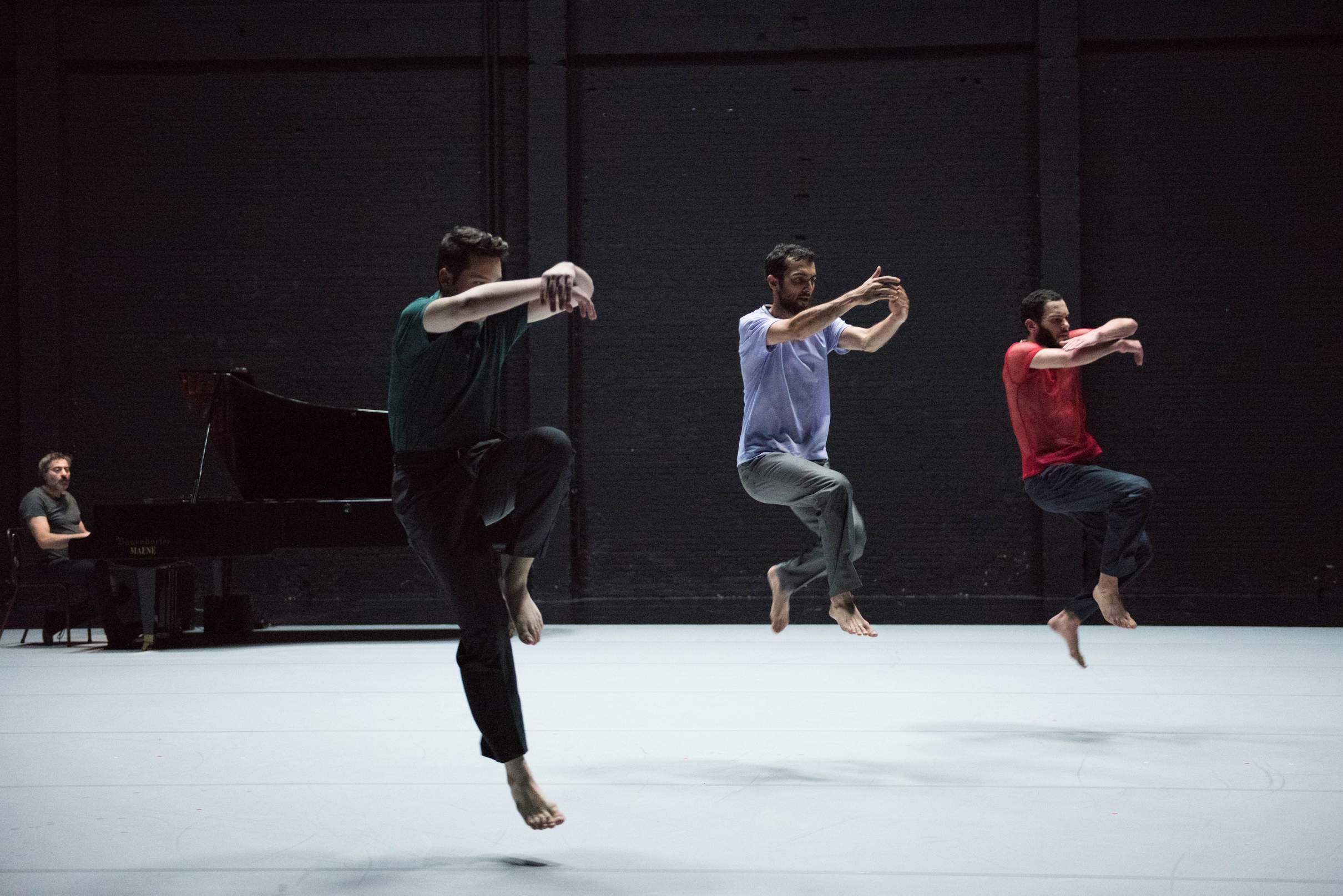 Three dancers are suspended mid-air, about to land from a unison jump, arms lifted to chest height, while a pianist watches from the back corner of the stage.