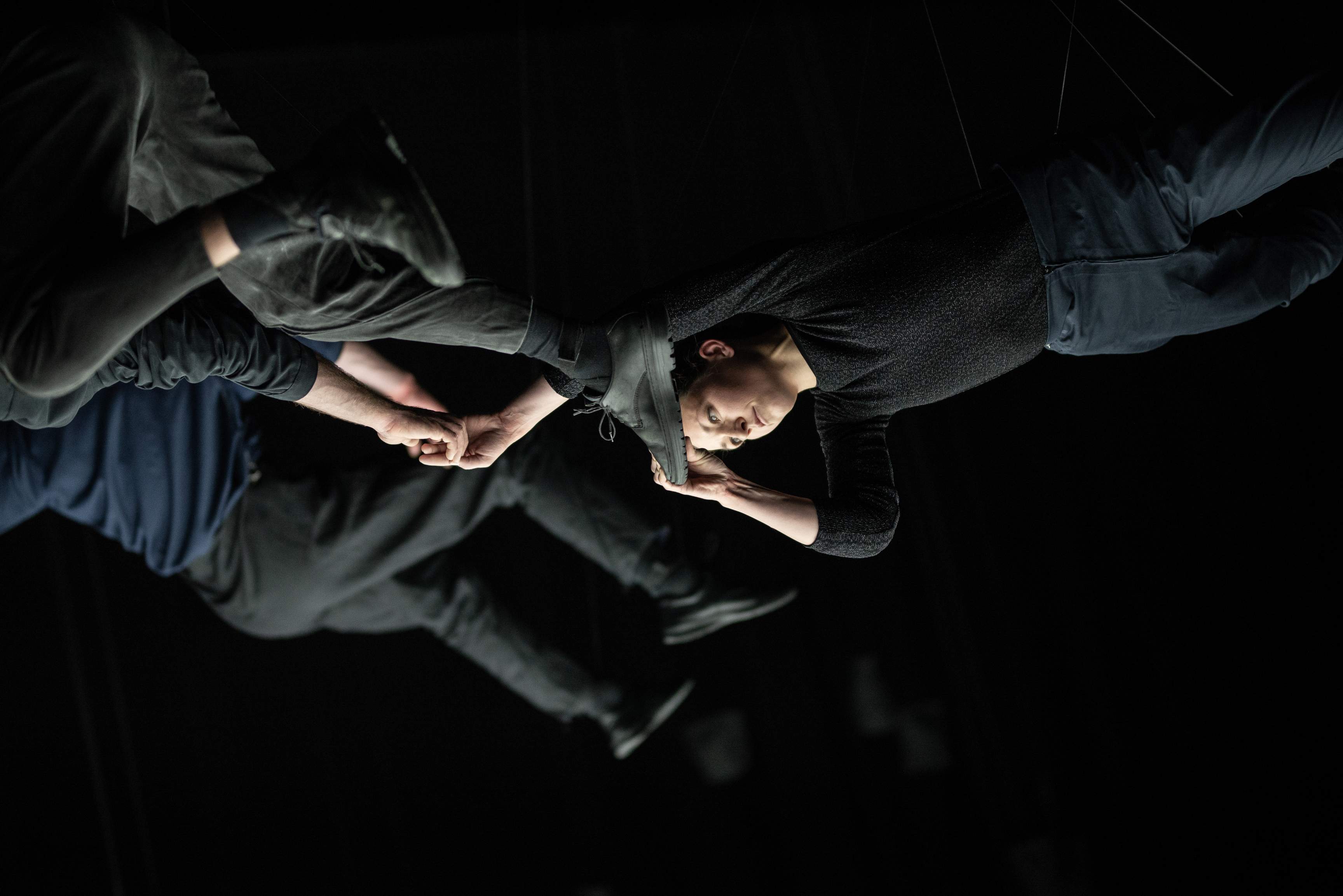 Three performers hang in midair by thin ropes in a dark space lit from below; two of them cling to each other by hands and feet.