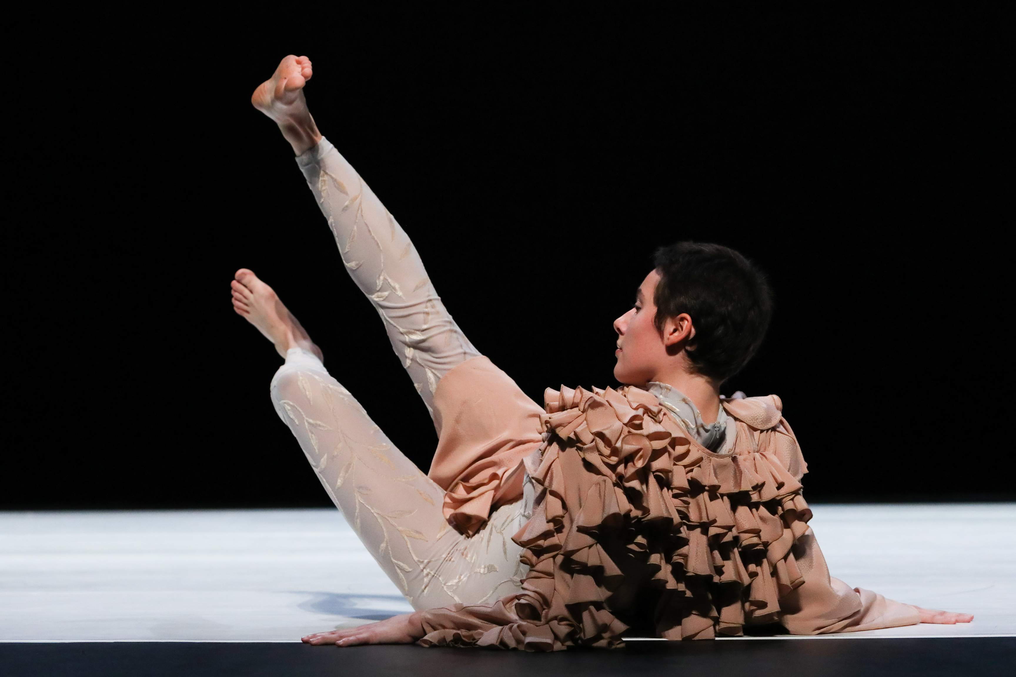 A young dancer wearing an elaborate costume lies on the floor during a dance phrase, legs stretched upward at an angle toward the ceiling.