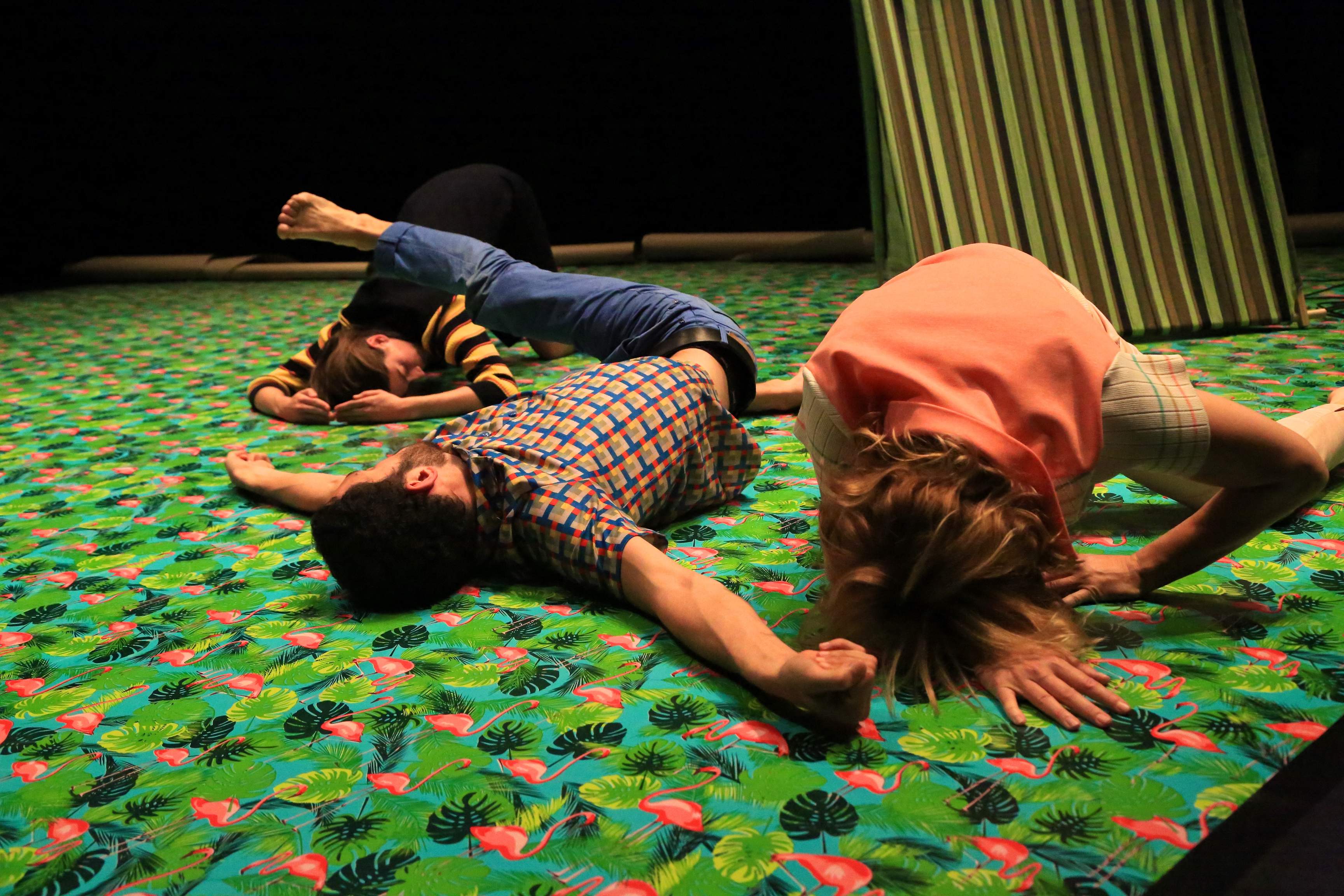Three performers lie on a brightly patterned floor, their bodies arranged to resemble a geographic landscape.