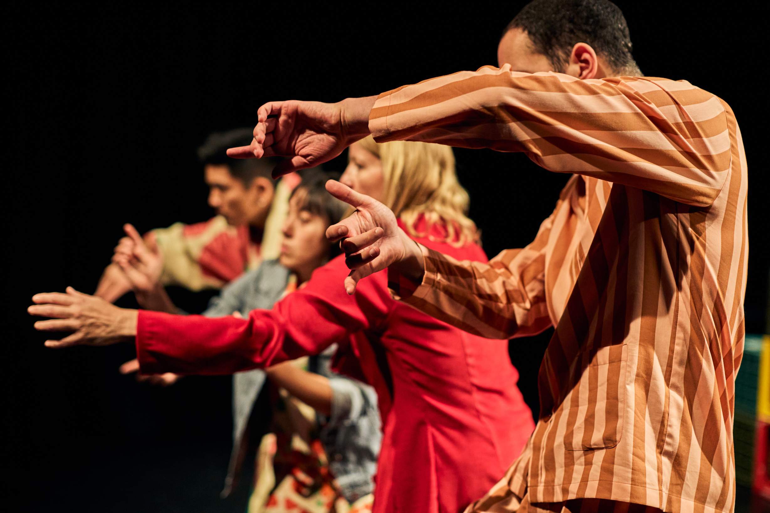 A line of performers in colourful outfits is seen from the side, making expressive hand gestures as they perform, resembling a secret or symbolic sign language.