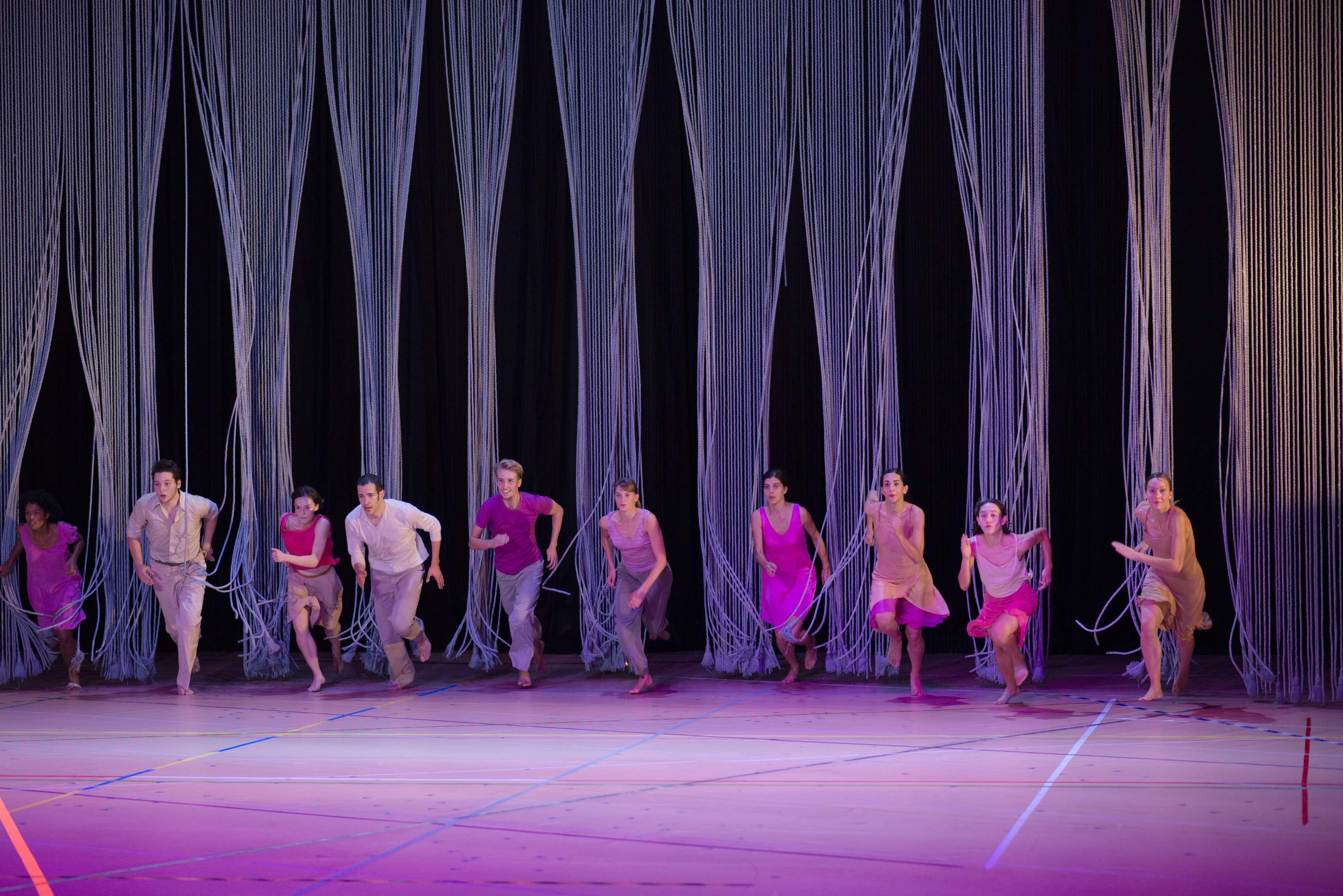 A large group of dancers races from the back to the front of the stage, moving through a curtain of ropes with eager enthusiasm.