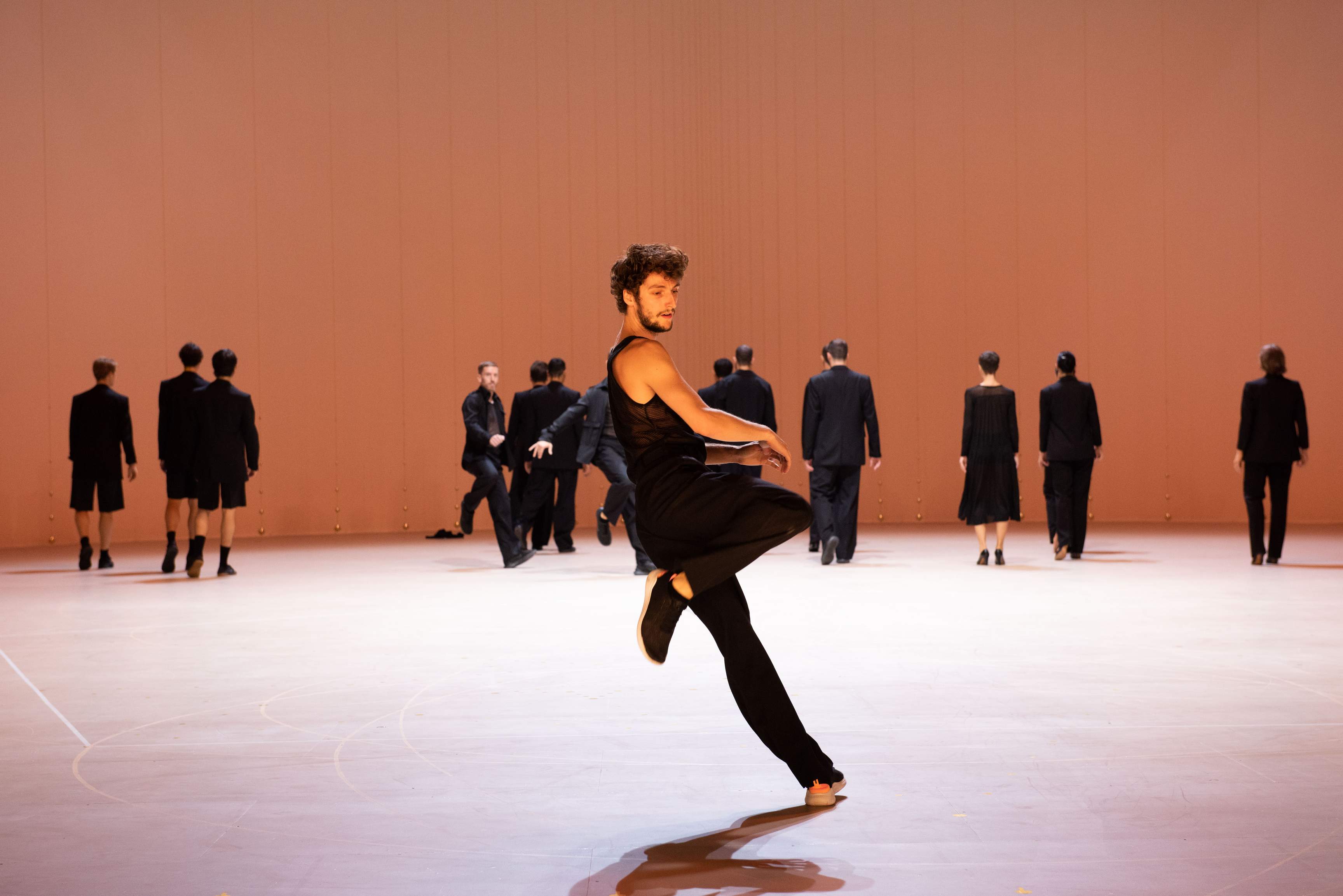 A dancer in the foreground pivots on his left leg, while several dancers walk backward at the back of the stage.