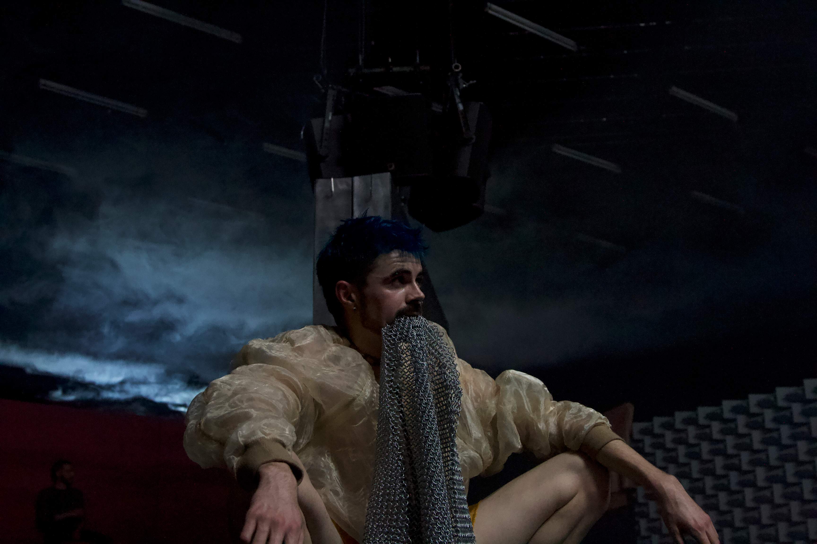 A performer crouches, holding a piece of chain mail between their teeth.