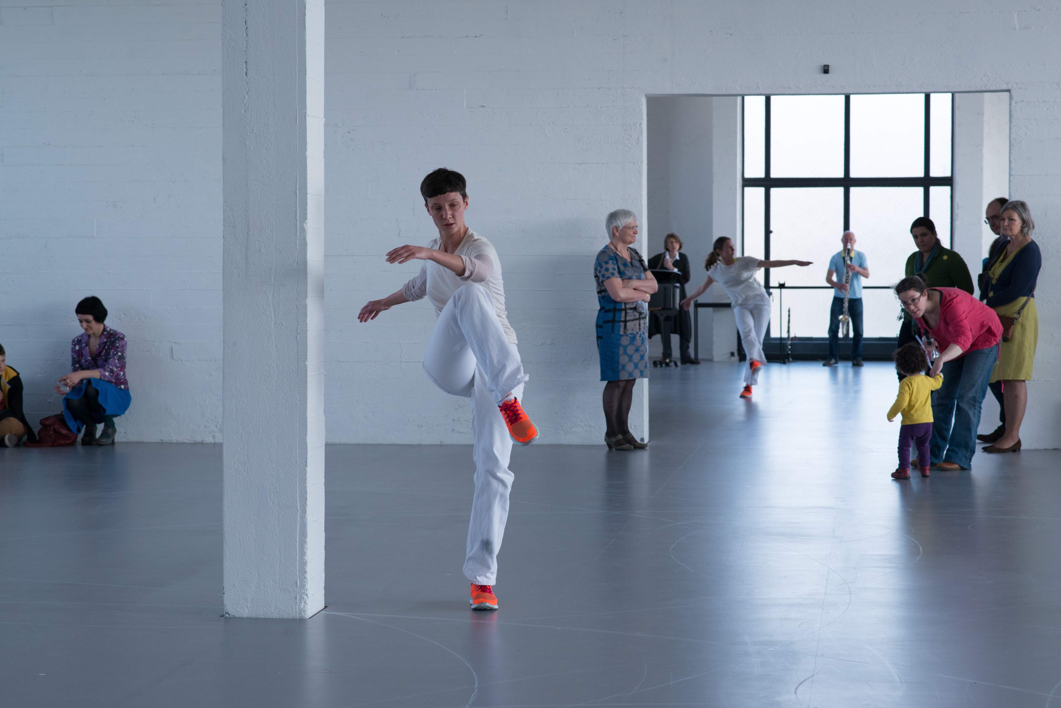 A dancer in a white museum space twists with one leg lifted, while another dancer and a clarinetist appear in the background.
