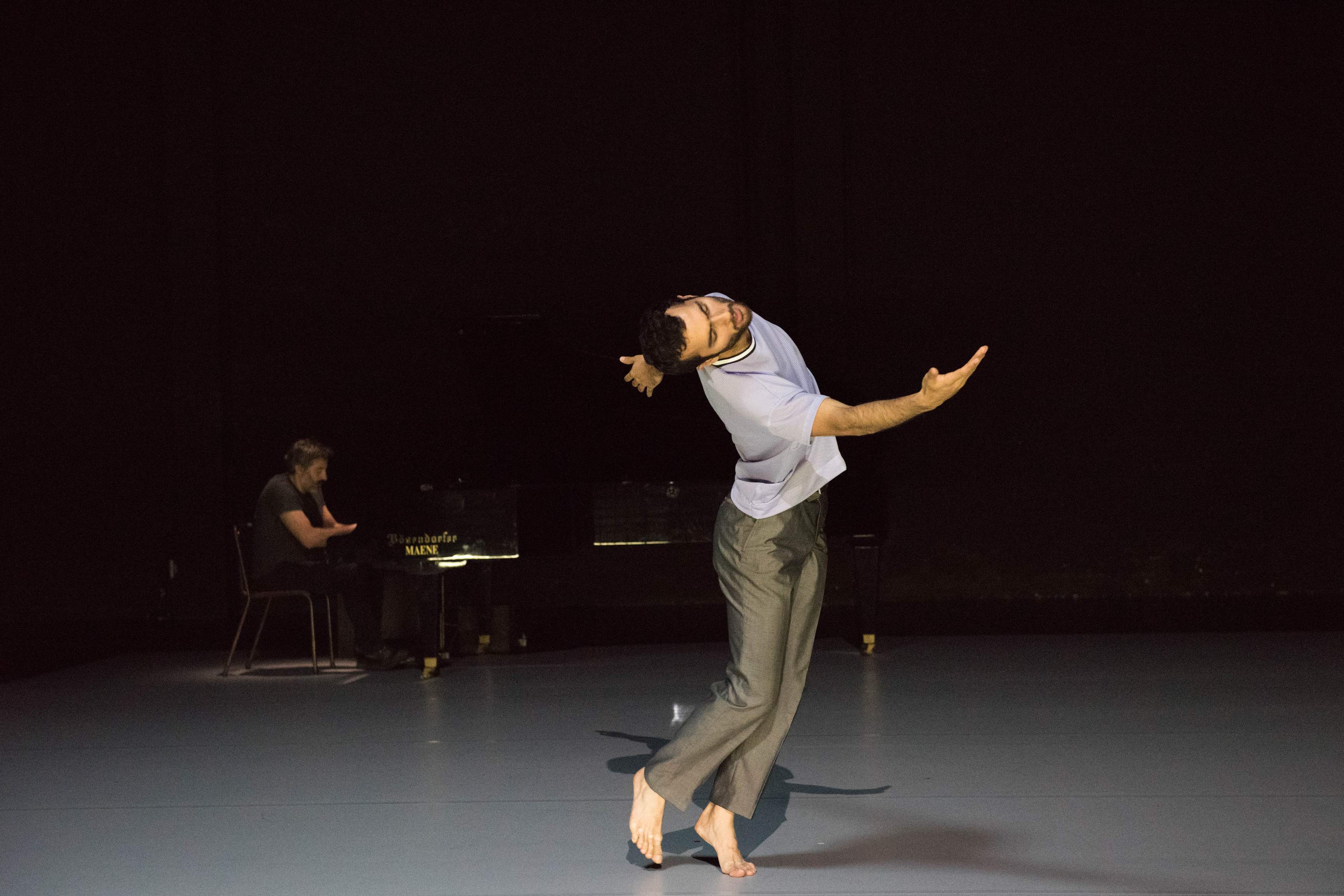 A dancer spirals the upper body, moving with a surrendering chest, while a pianist plays behind him.